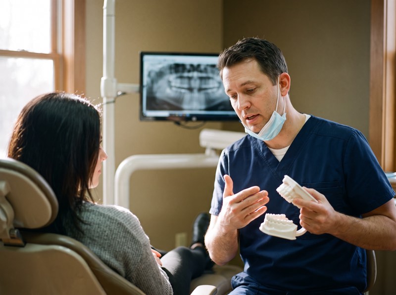 A dentist explaining a treatment plan to a patient using a model of the jaw, with an x-ray on screen behind
